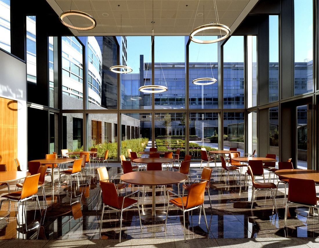 ABN AMRO: Double-height dining hall with orange chairs, round tables and ring-shaped pendant lights in front of a glass wall