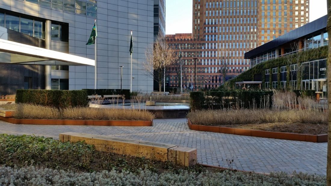 ABN AMRO: Paved garden with raised planting beds and wooden benches beside glass buildings and flags