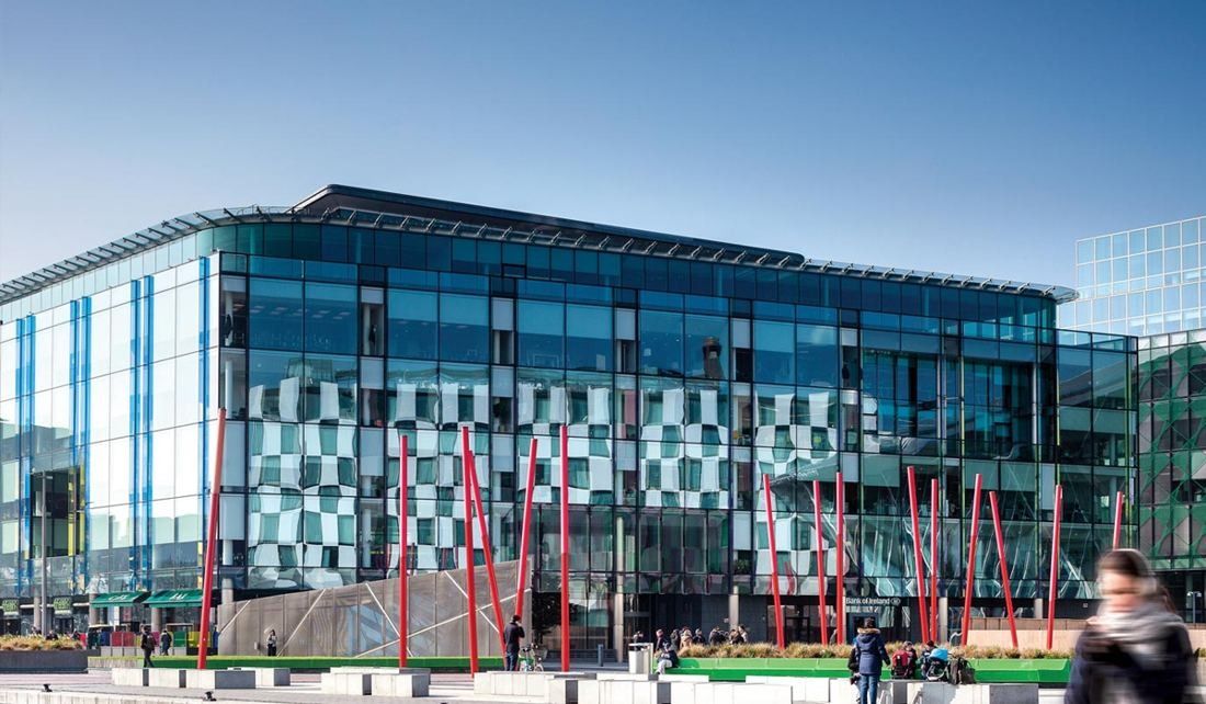 Accenture: Glass facade of a five-storey office building reflecting a checkered building opposite, red poles on the square in front
