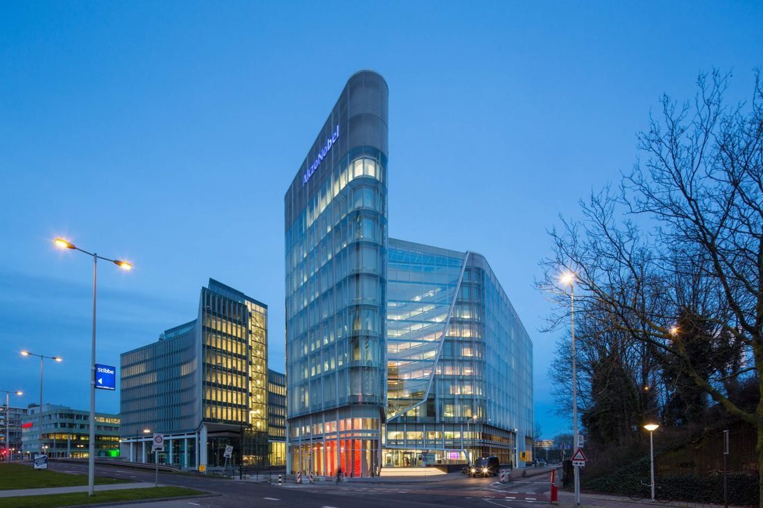 Curved glass office tower and lower glass wing lit at dusk beside a road with street lamps