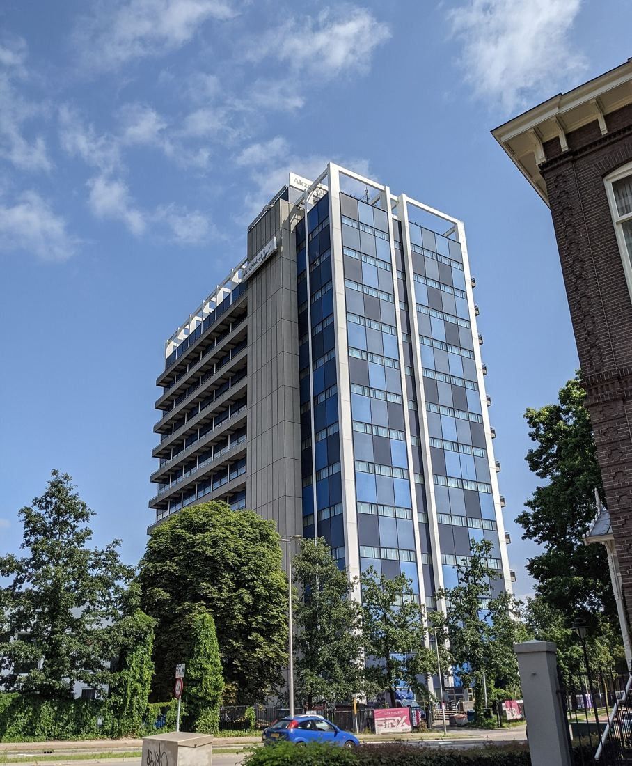 Tall office building with blue window bands behind trees, next to an older brick building