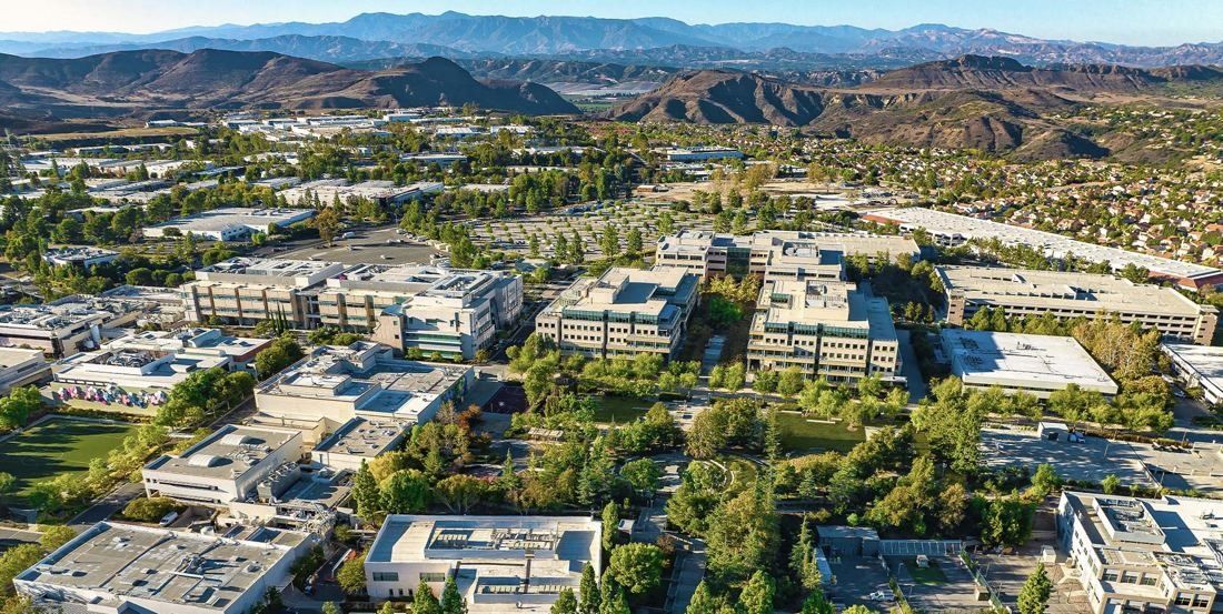 Aerial view of a large campus of low white buildings among trees, with mountains on the horizon