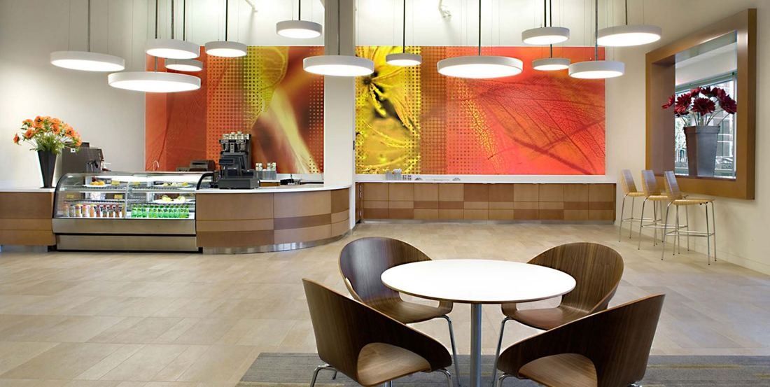 Amgen: Café with round white tables, curved chairs, pendant lights and large orange and yellow artwork behind the counter