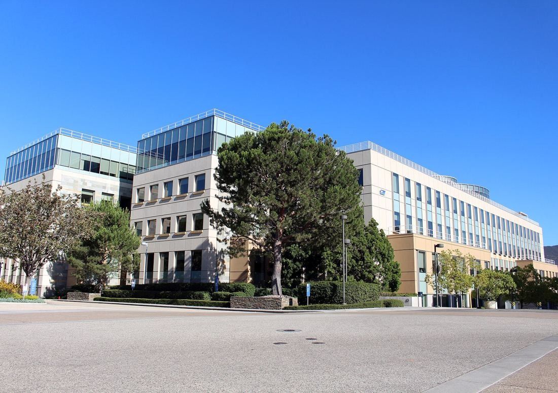 Amgen: Four-storey office buildings with glass and stone facades behind trees and a wide empty paved area