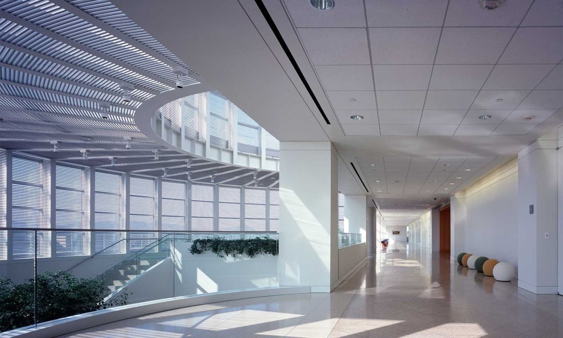 Amgen: Long white corridor beside a glass wall with louvres, looking over a lower-level atrium