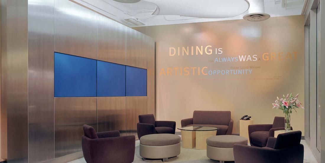 Amgen: Lounge with brown armchairs and round ottomans beside a metal wall with three screens and wall lettering