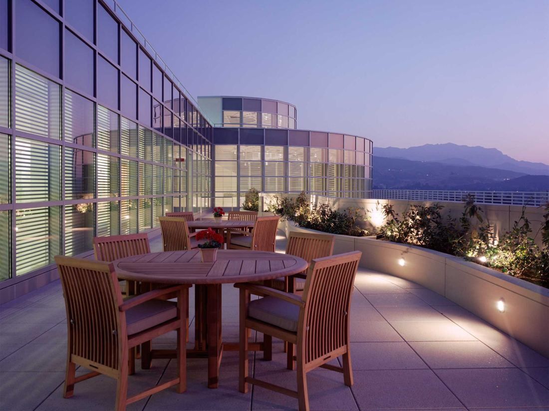 Amgen: Terrace with wooden tables and chairs at dusk beside a glass facade, mountains in the distance