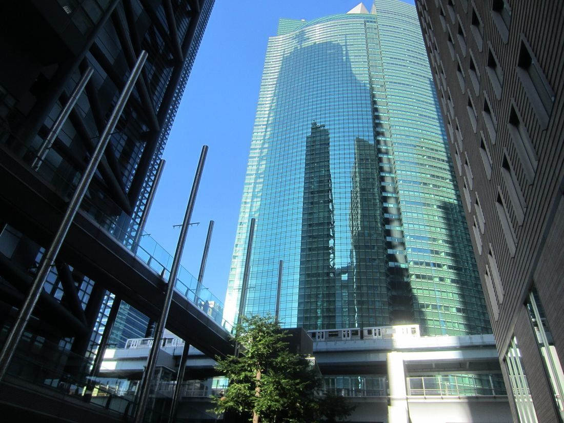 Curved green glass tower rising above a raised walkway and street trees under a blue sky