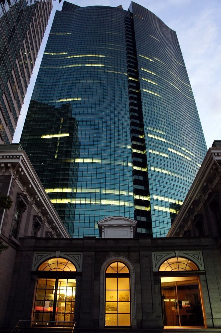 ANA: Glass tower at dusk rising behind a low historic building with lit arched windows