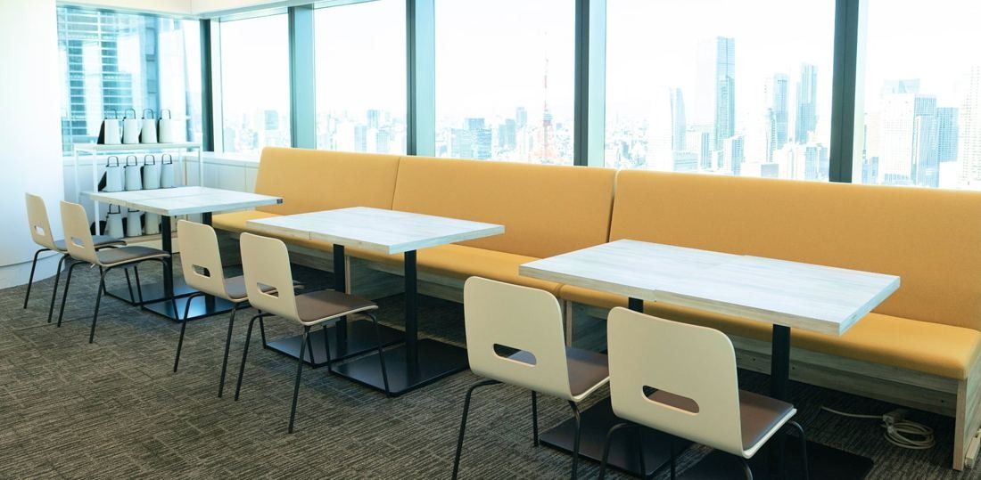 ANA: Long orange bench with white tables and chairs along floor-to-ceiling windows overlooking the city