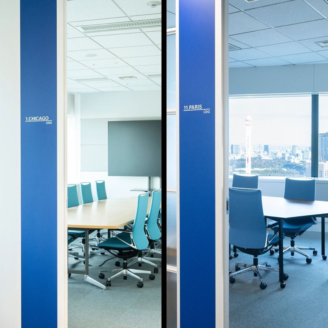 ANA: Meeting room seen through glass doors with blue panels, a table, blue chairs and a city view