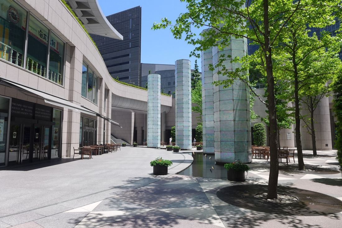 ANA: Paved open plaza with trees and tall glass light columns beside the podium