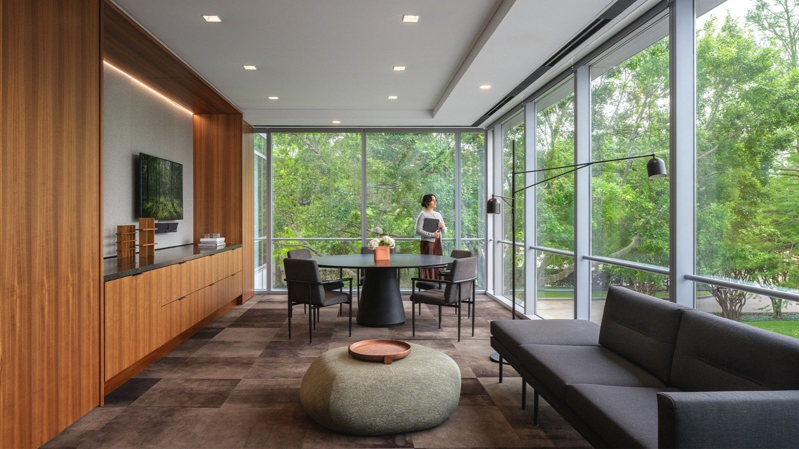 A lounge with a long dark sofa and a round table beside windows full of trees