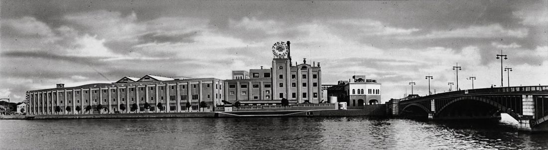 Black-and-white panorama of long brewery buildings with a clock tower on the riverbank next to an arched bridge