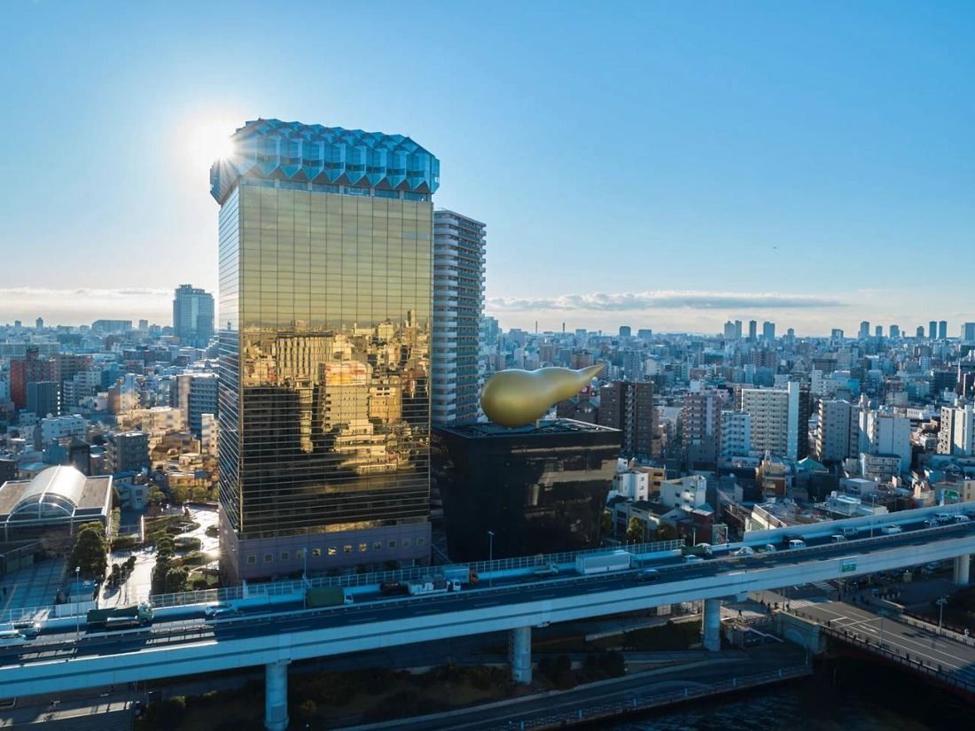 Gold mirrored-glass tower with a white crown beside a black hall topped by a golden flame, seen from above the river in sunlight