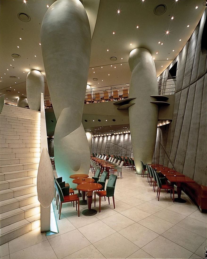 Asahi Group Holdings: Tall hall interior with huge white bulbous columns, a lit white stone staircase and rows of wooden tables with green chairs