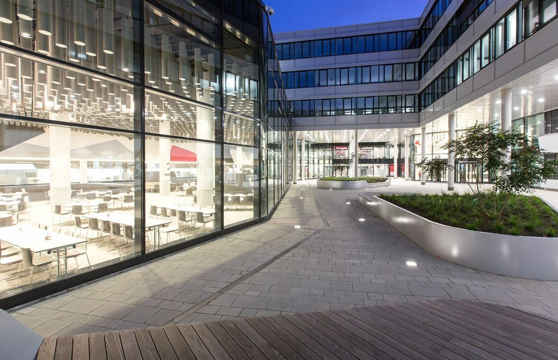 Audi: Glass-walled canteen with rows of tables beside a paved courtyard with planters, at dusk