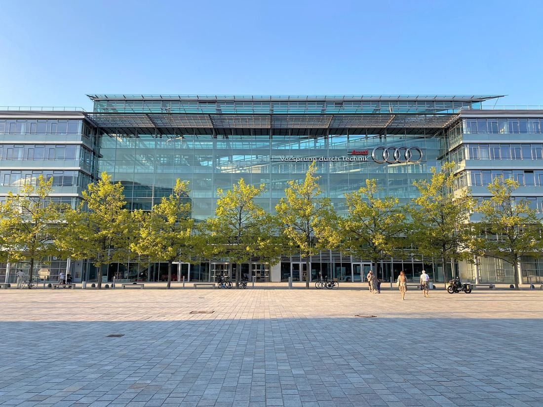 Audi: Large glass-fronted office building behind a row of young trees and a wide paved square