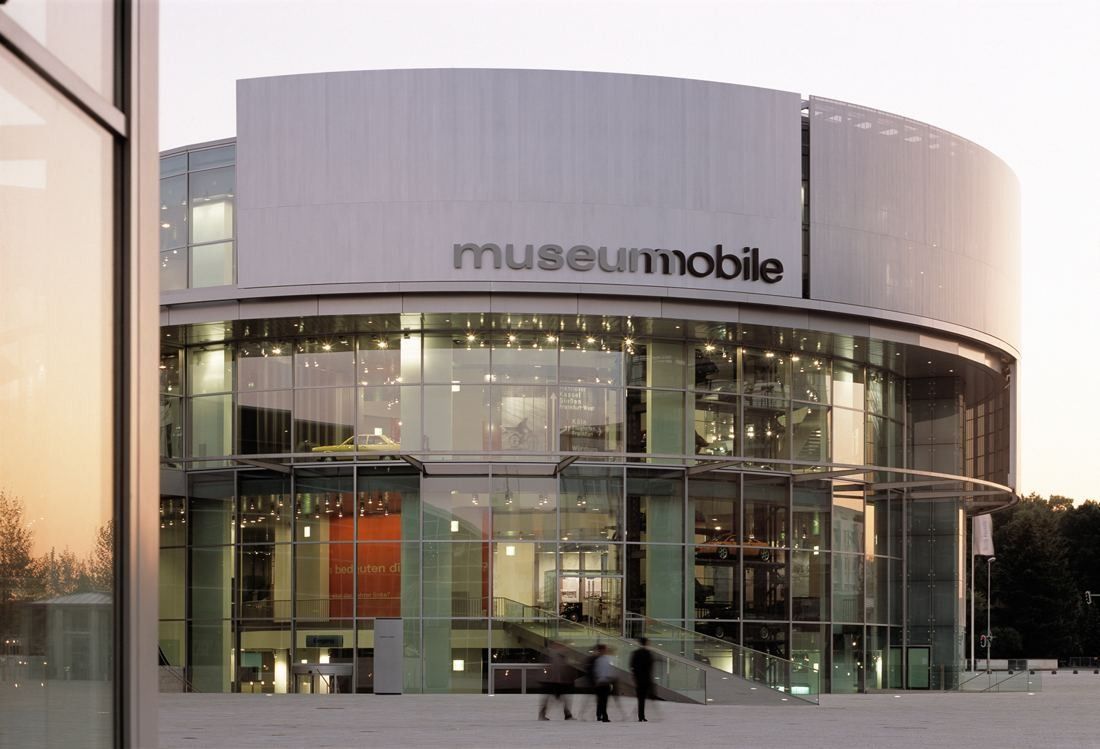 Round glass building with a silver band reading museum mobile, lit from inside at dusk