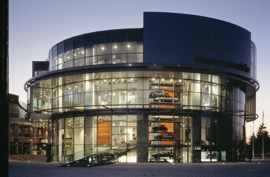 Audi: Round glazed building lit at dusk, with cars visible on platforms behind the glass
