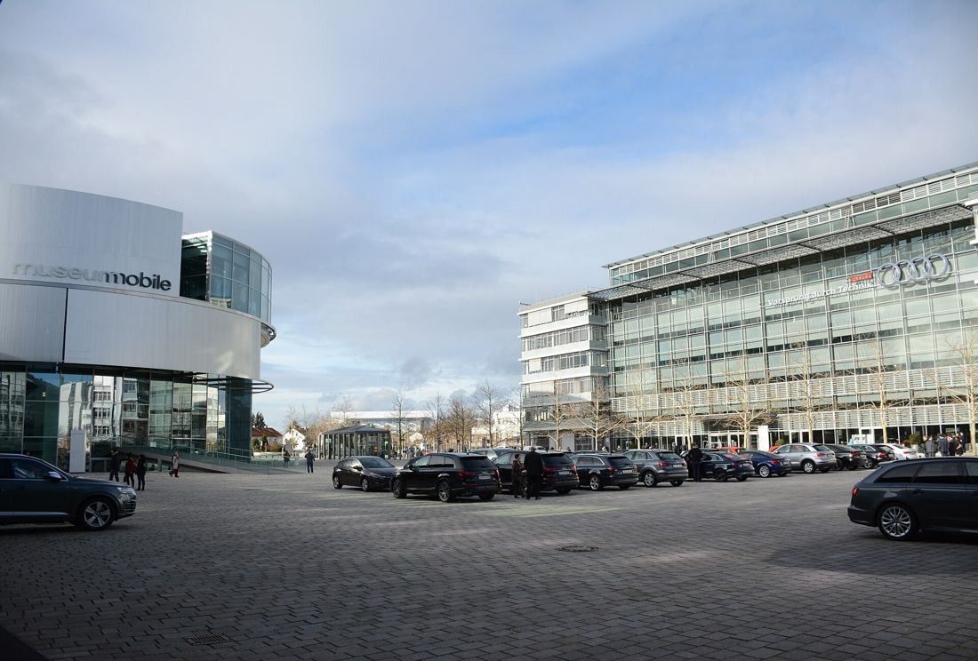Audi: Round museum building beside a glass office block and a car park under a cloudy sky