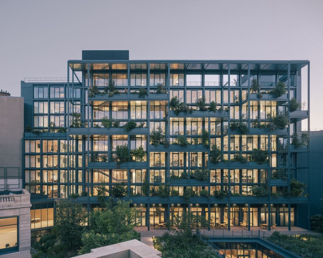 Glass-and-steel office building with planted terraces on every level, lit at dusk above a garden