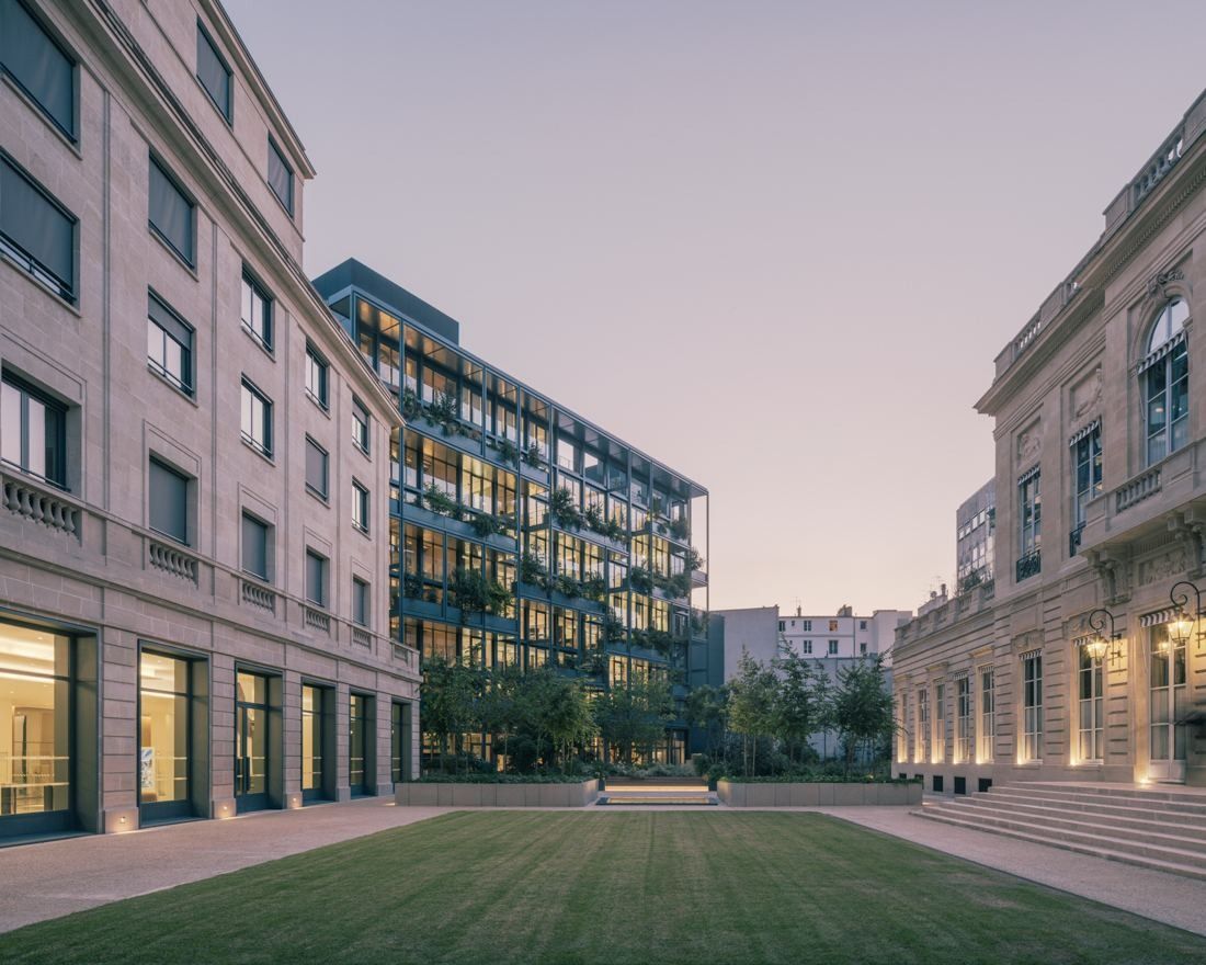 AXA: Lawn courtyard framed by stone façades and a glazed, planted office building at dusk