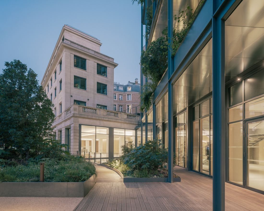 AXA: Wooden deck and planting between a stone building and a glazed façade at dusk