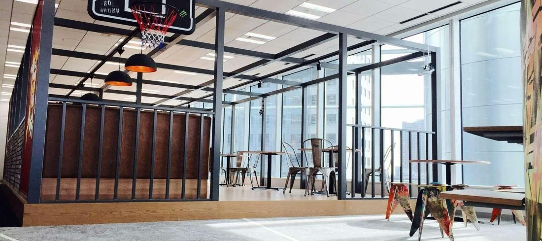Bandai Namco: Glass-walled corner room with a basketball hoop, wooden slats and cafe tables by tall windows