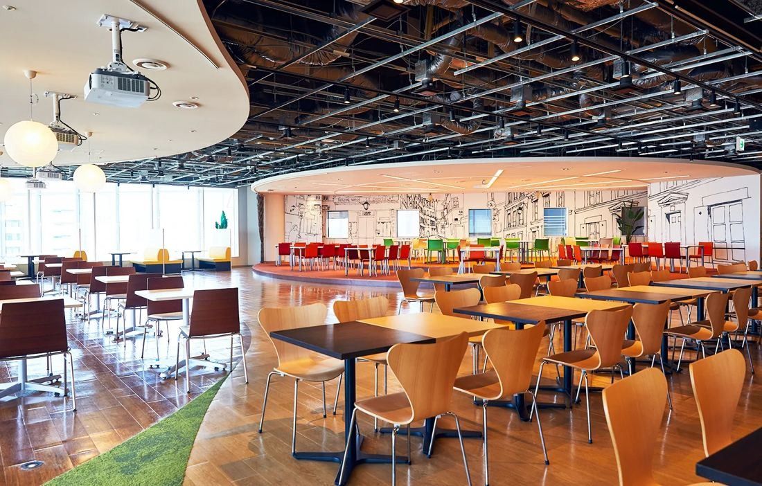 Bandai Namco: Large cafeteria with wooden chairs, round tables and a colourful mural under an exposed ceiling