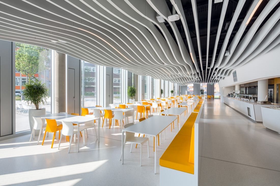 Bright canteen with long white tables, orange chairs and a wave-shaped slatted ceiling