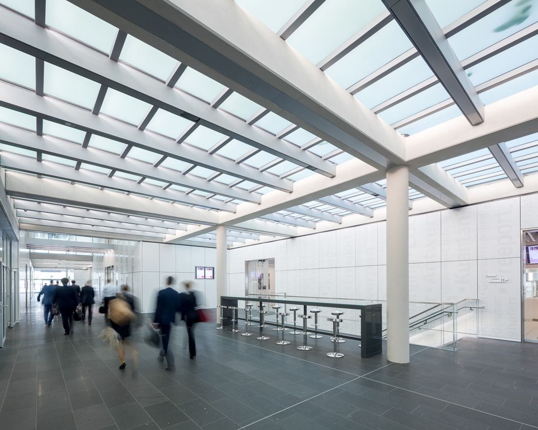 Long interior street under a glazed roof with white beams, people walking past a standing bar