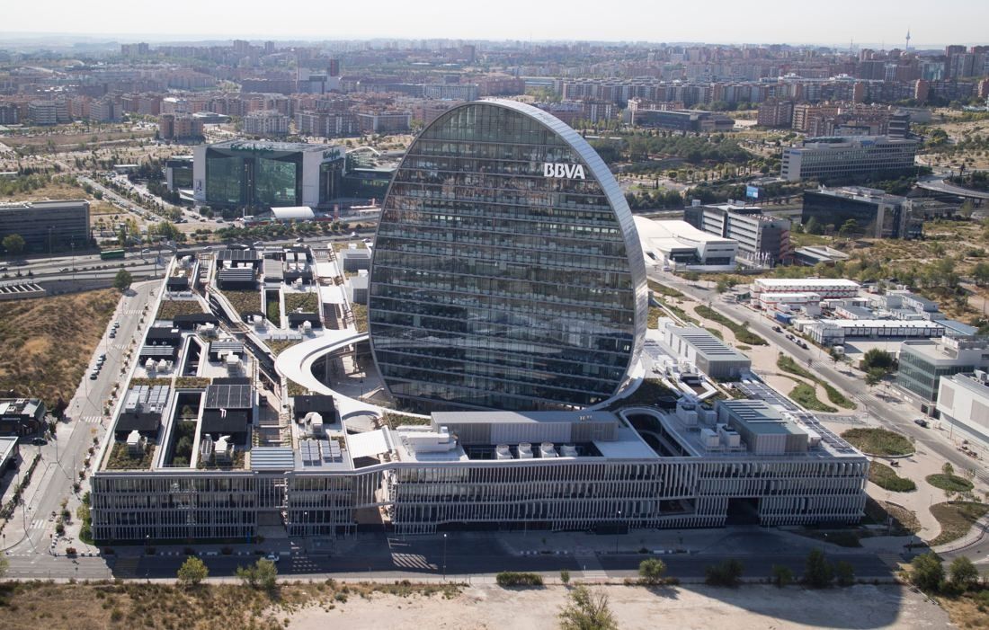 Aerial view of an oval glass tower rising from a large low-rise office complex with roof gardens, surrounded by roads and a city district