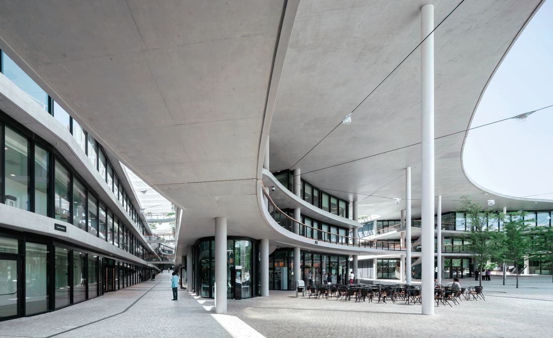 BBVA: Covered walkway under a curving concrete canopy beside glazed office wings, with outdoor tables