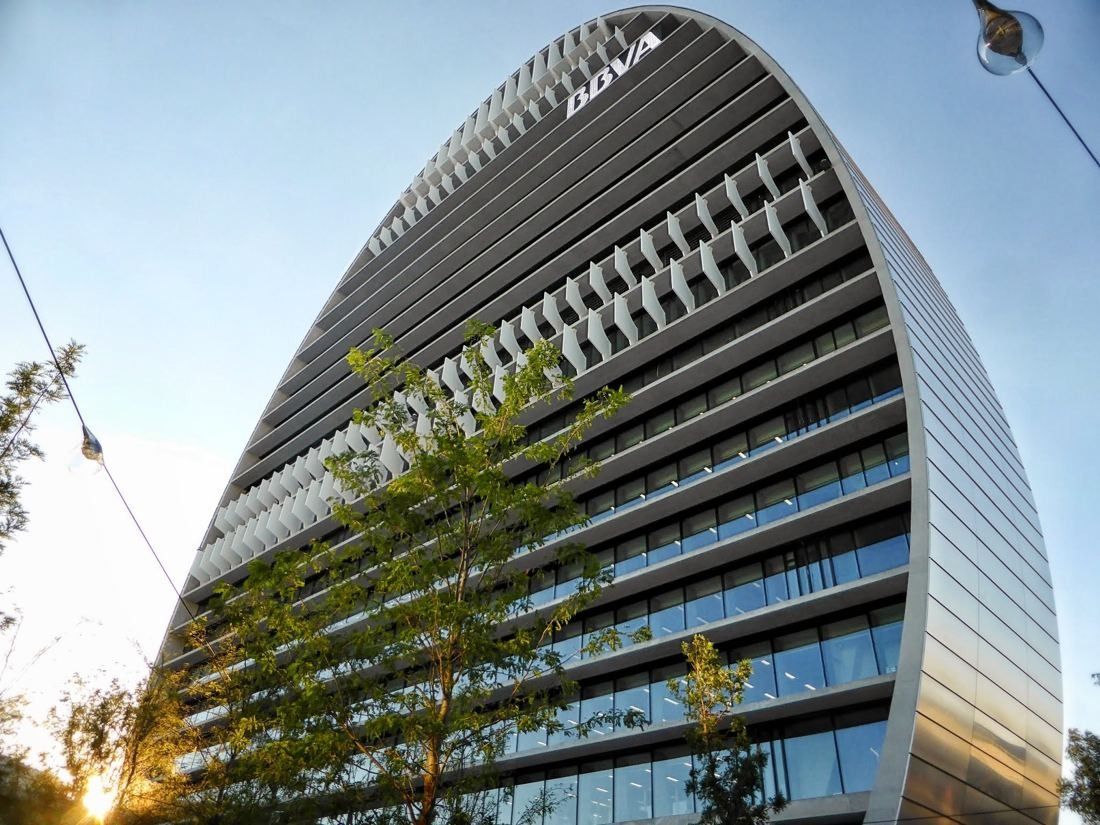 BBVA: Curved tower façade with horizontal shading fins seen from below beside a young tree