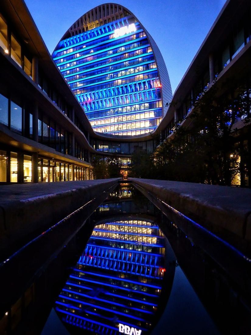 BBVA: Illuminated oval tower at night reflected in a long water channel between buildings