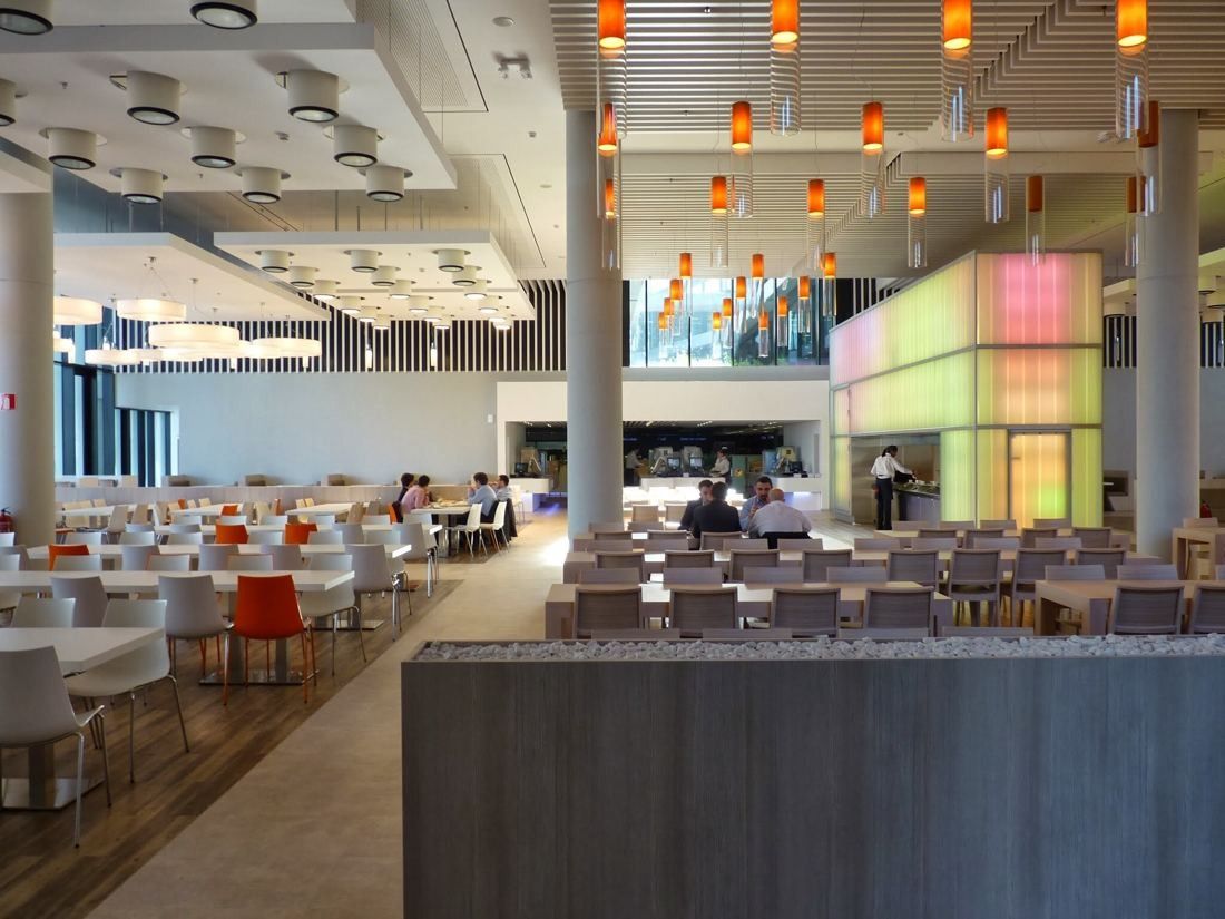 BBVA: Large canteen with long tables, orange pendant lights and a glowing glass kiosk