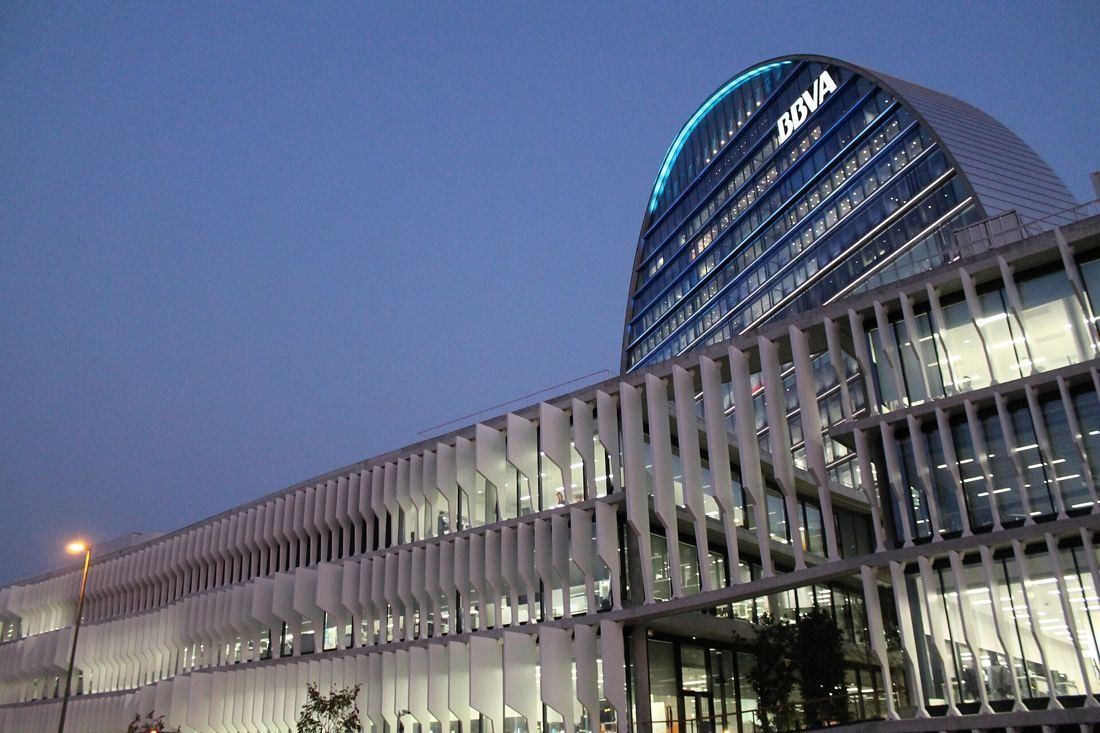 BBVA: Lit oval tower and a long low-rise façade of white fins at dusk