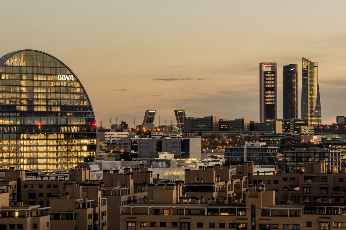 BBVA: Lit oval tower at dusk with a row of skyscrapers in the distance