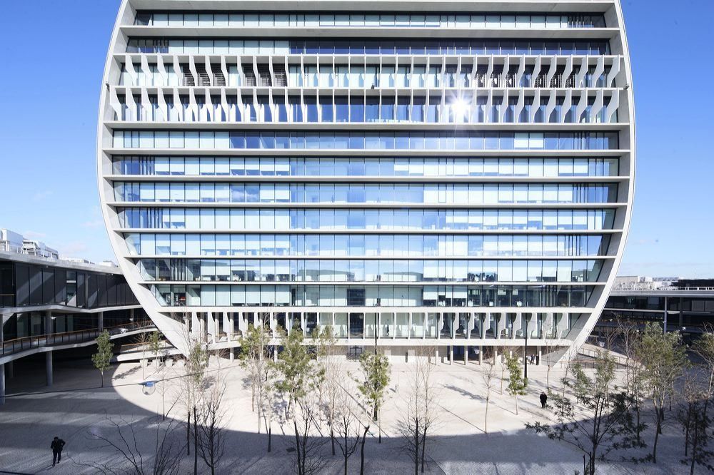 BBVA: Oval tower with horizontal bands of glass and shading fins above a paved plaza planted with young trees