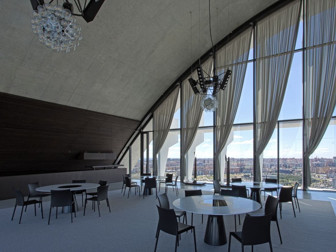 BBVA: Room with round tables under a sloping concrete ceiling and tall curtained windows over the city