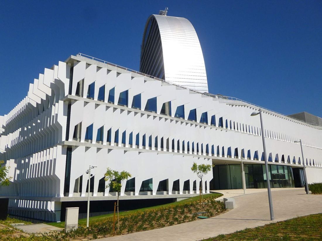 BBVA: White-finned low-rise building with a landscaped path and the curved top of the tower behind