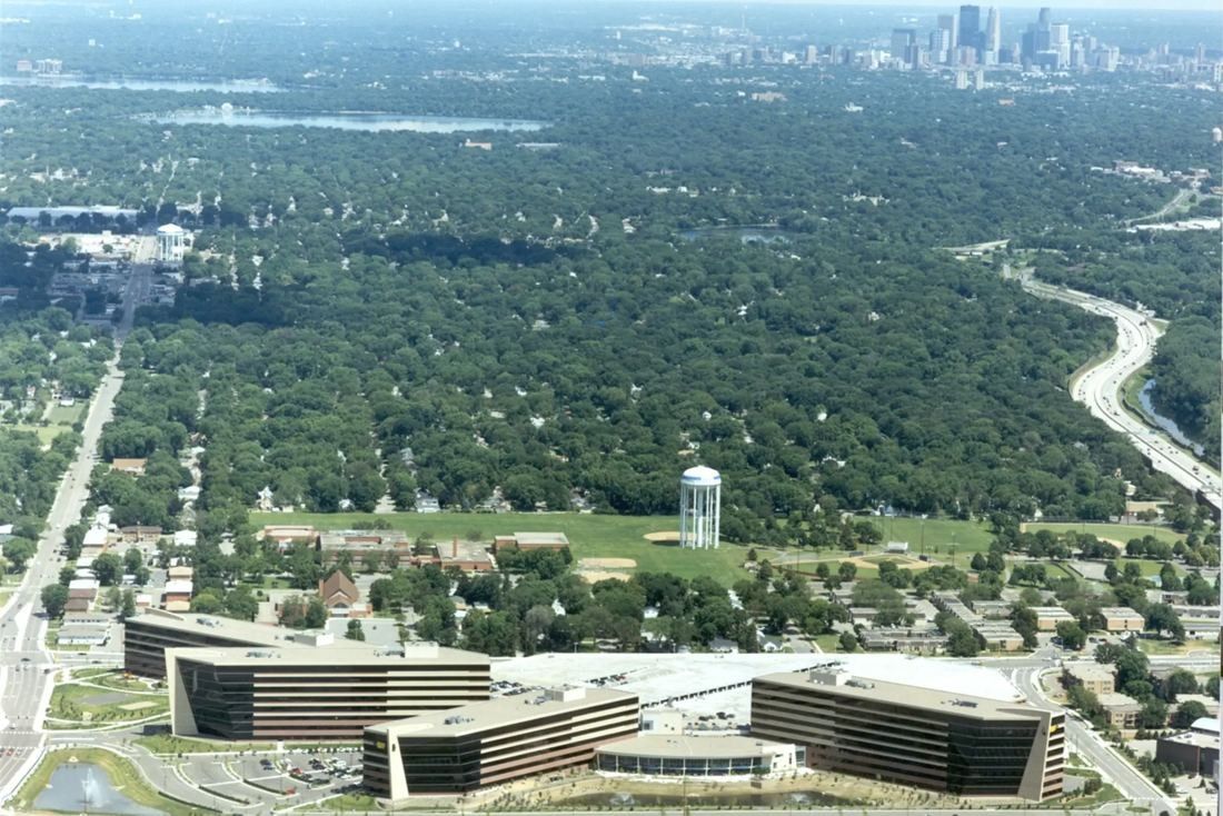 Aerial view of a large office campus of several low-rise and mid-rise buildings beside a highway, with trees and a city skyline in the distance