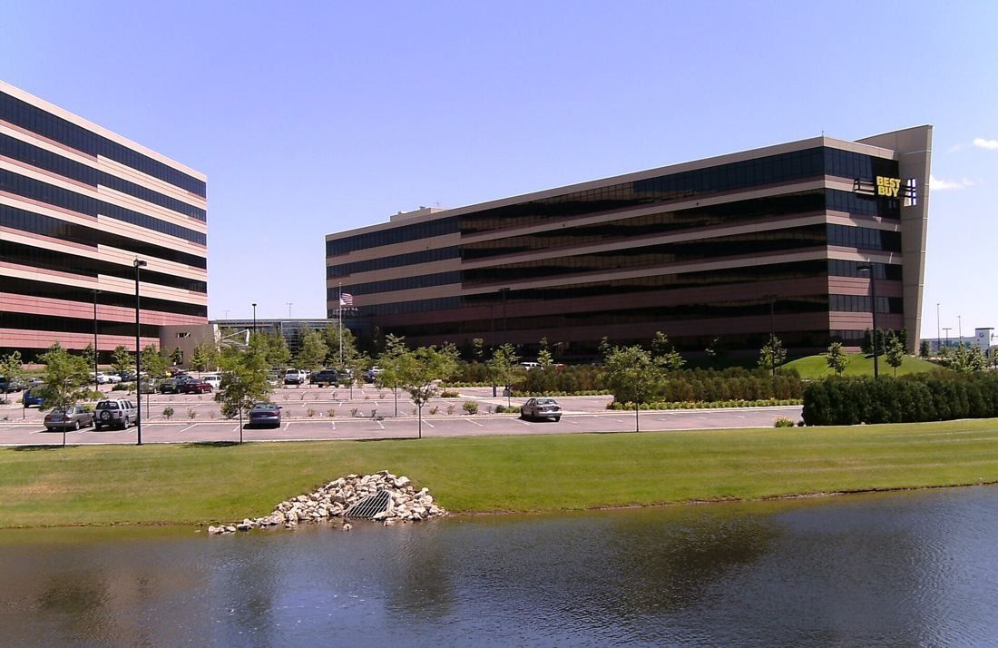 Best Buy: Dark banded office buildings behind a pond, lawn and parking lot on a sunny day