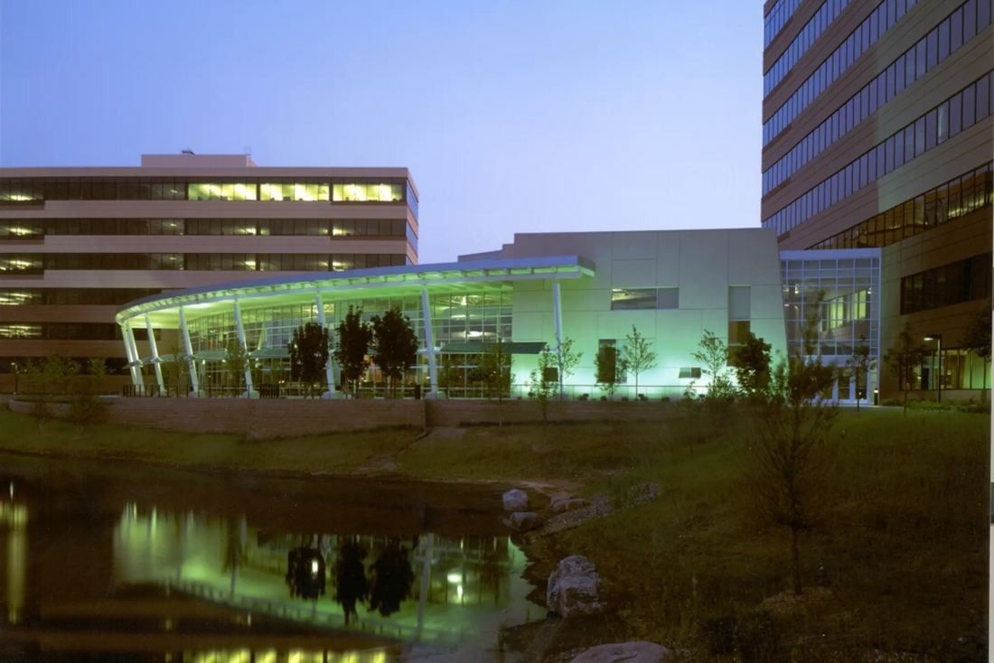 Best Buy: Glass-fronted low building lit green at dusk, reflected in a pond between taller office blocks