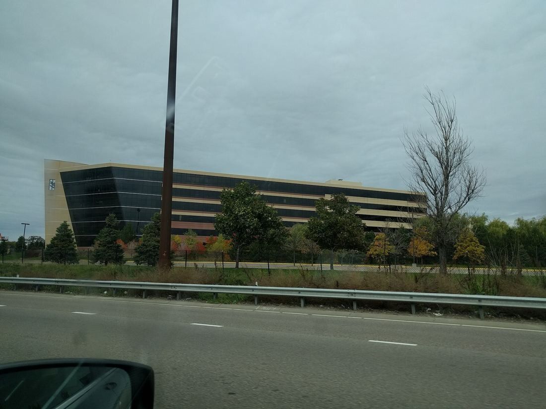 Best Buy: Long office building seen across a highway from a passing car, autumn trees in front