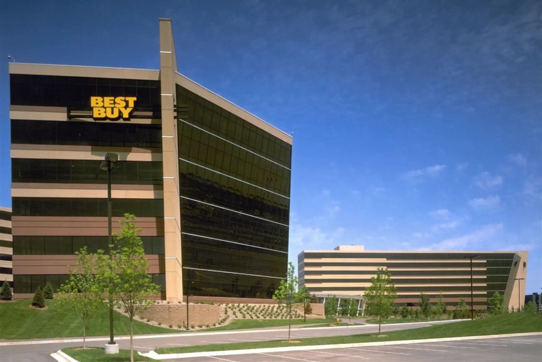 Best Buy: Tall office tower with dark glass and a sign on top, with a lower building and young trees beside it