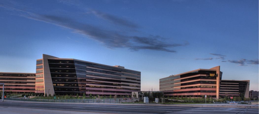 Best Buy: Wide view of angular office buildings under an evening sky across an open paved area