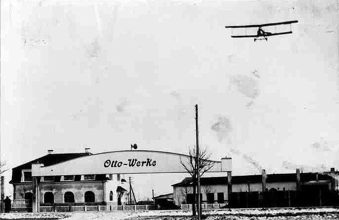 Low factory building with an Otto-Werke sign and a biplane overhead