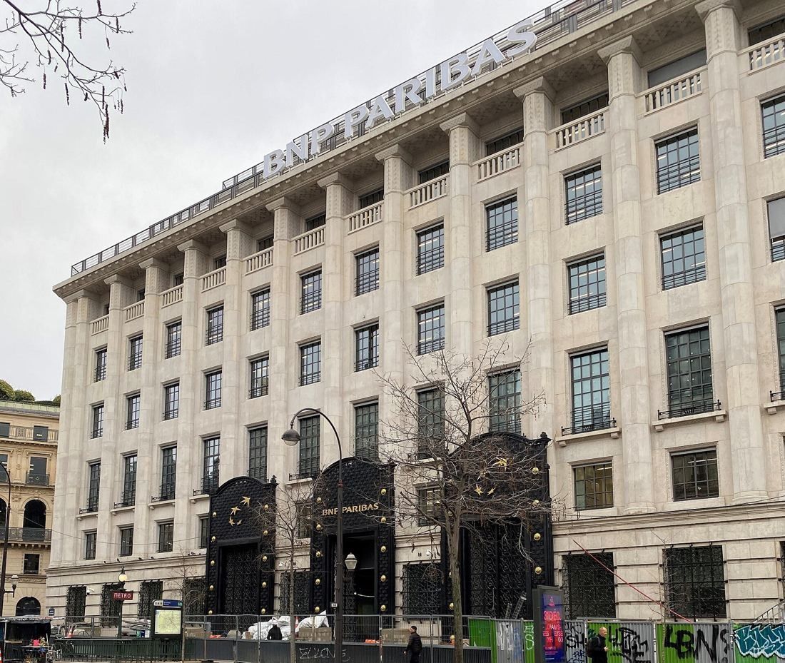 BNP Paribas: Stone facade with tall pilasters, a BNP Paribas sign and dark ornamental doors behind a site hoarding